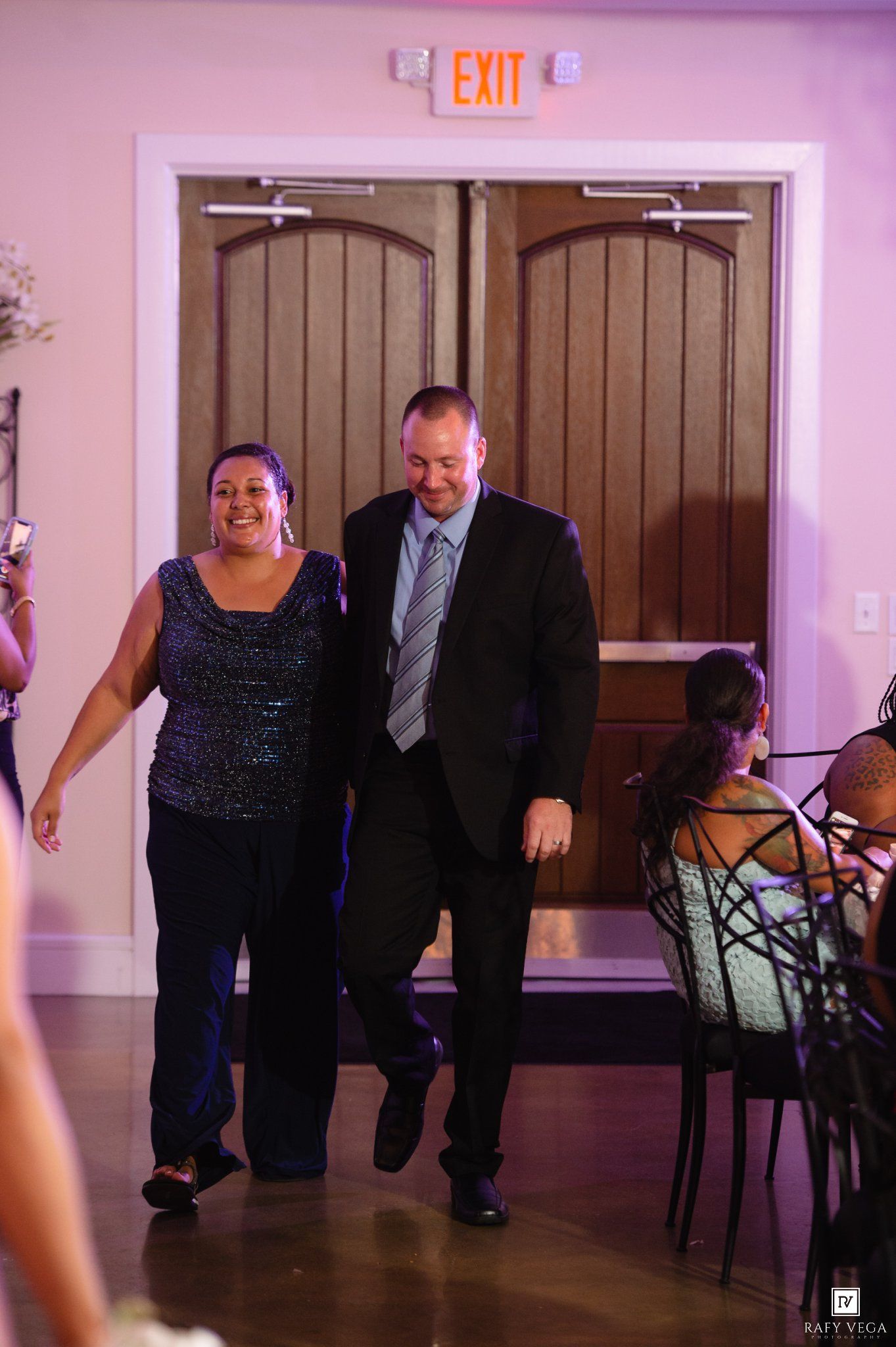A man in a suit and woman in sequin top walk through wooden doors at a wedding reception venue.