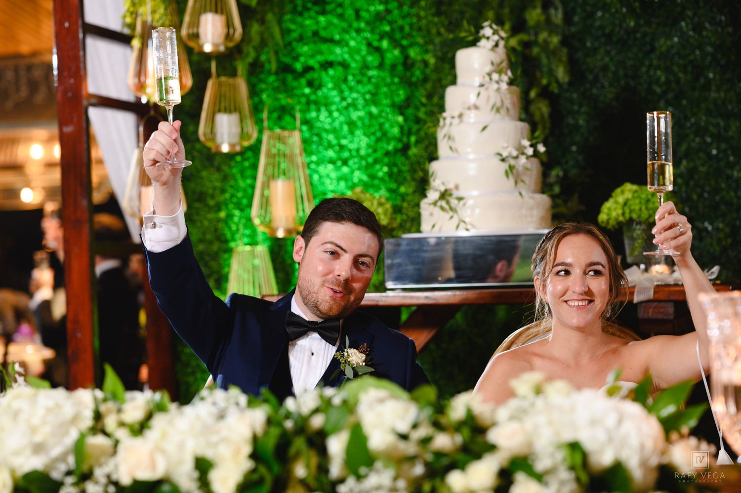 Newlyweds toast with champagne at wedding reception, white tiered cake and green floral wall backdrop.
