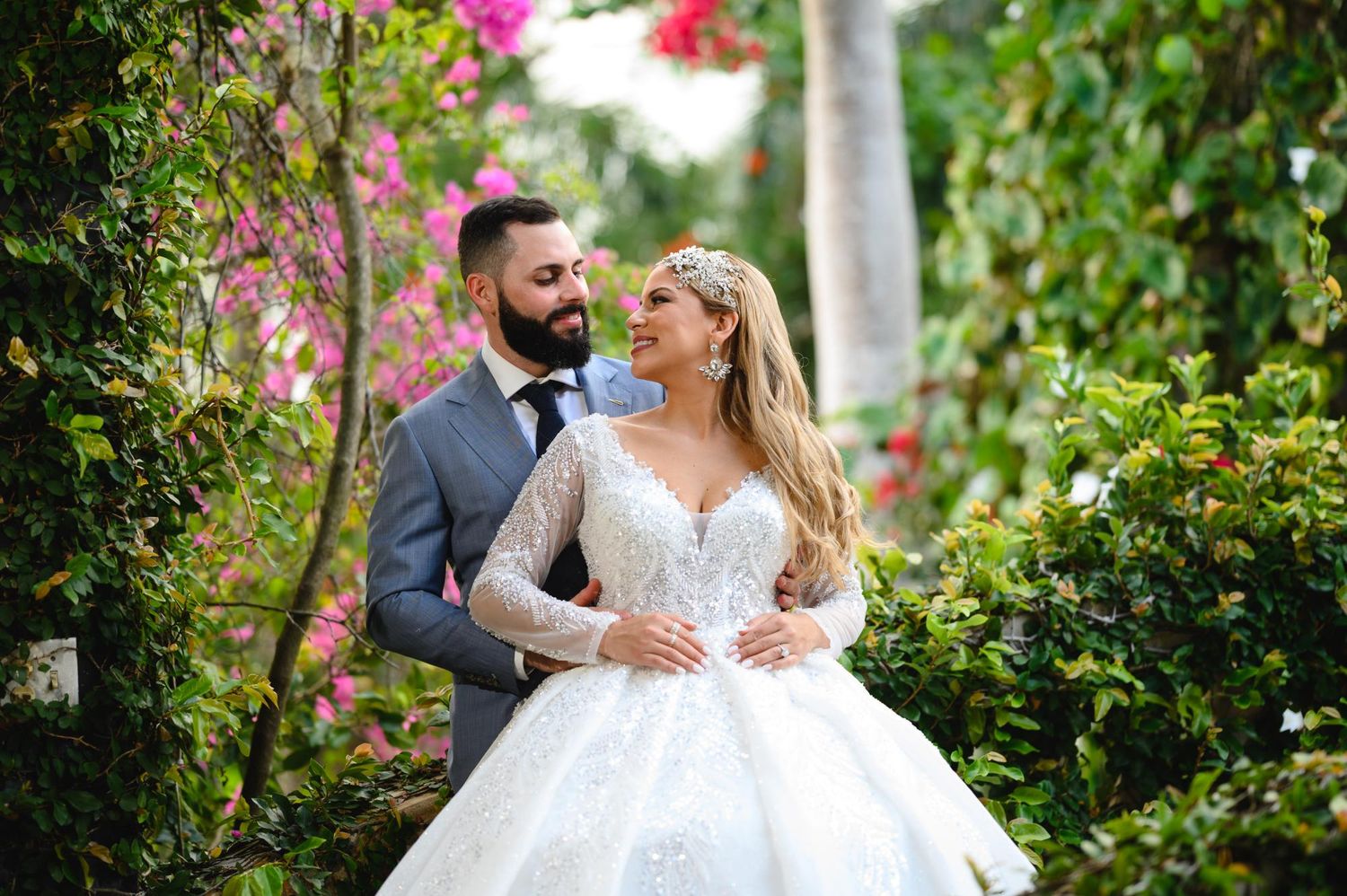 Bride in lace ball gown and groom in gray suit pose among lush greenery and pink flowers.