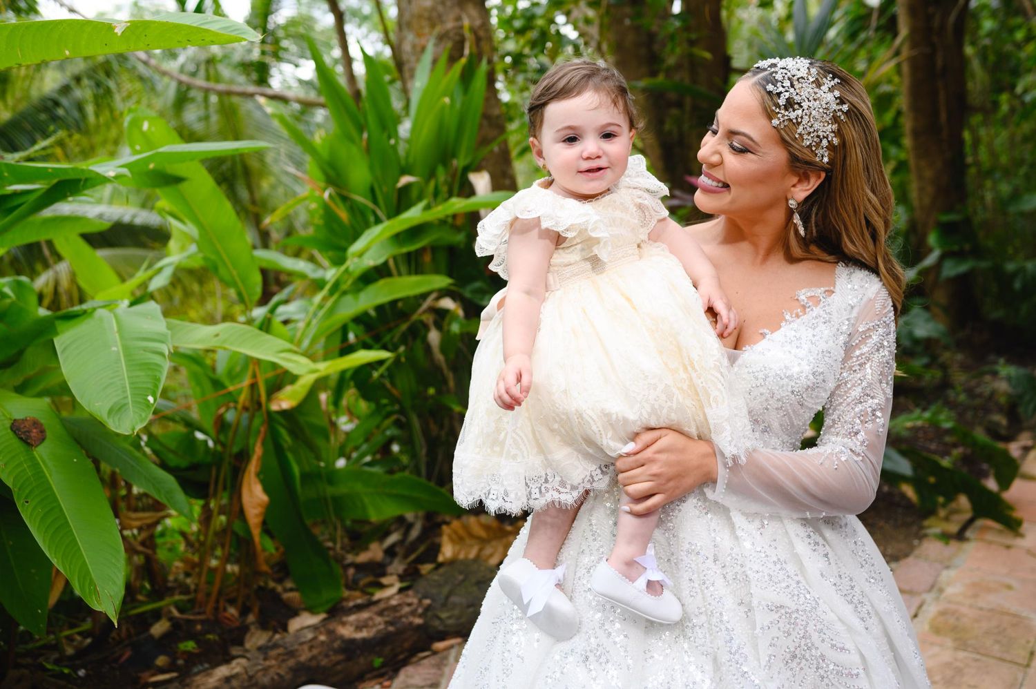 Bride in lace wedding gown holds smiling baby girl in white dress amid lush tropical greenery.