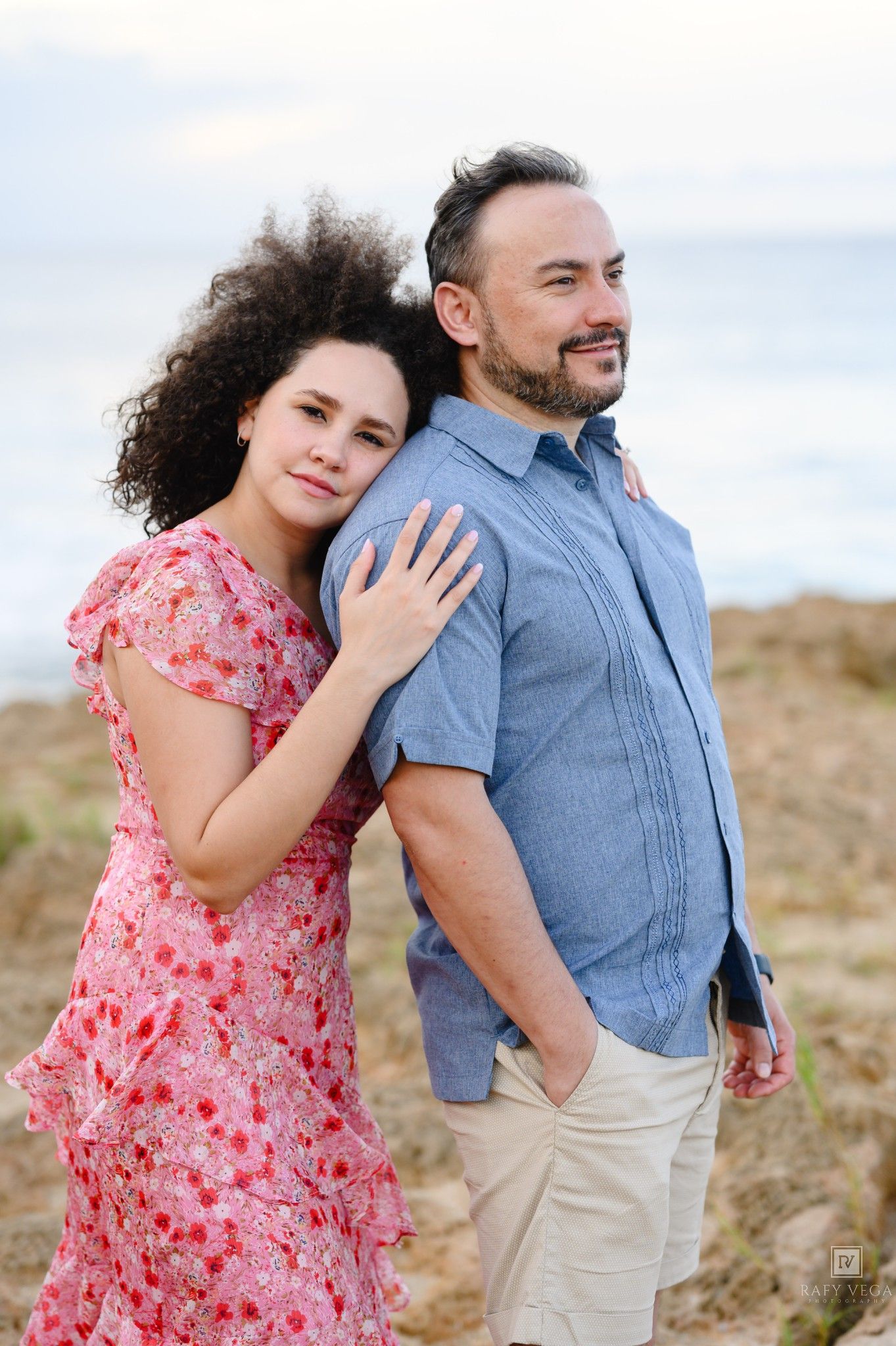 A woman in a pink floral dress and a man in a blue shirt pose back-to-back on a sandy beach.