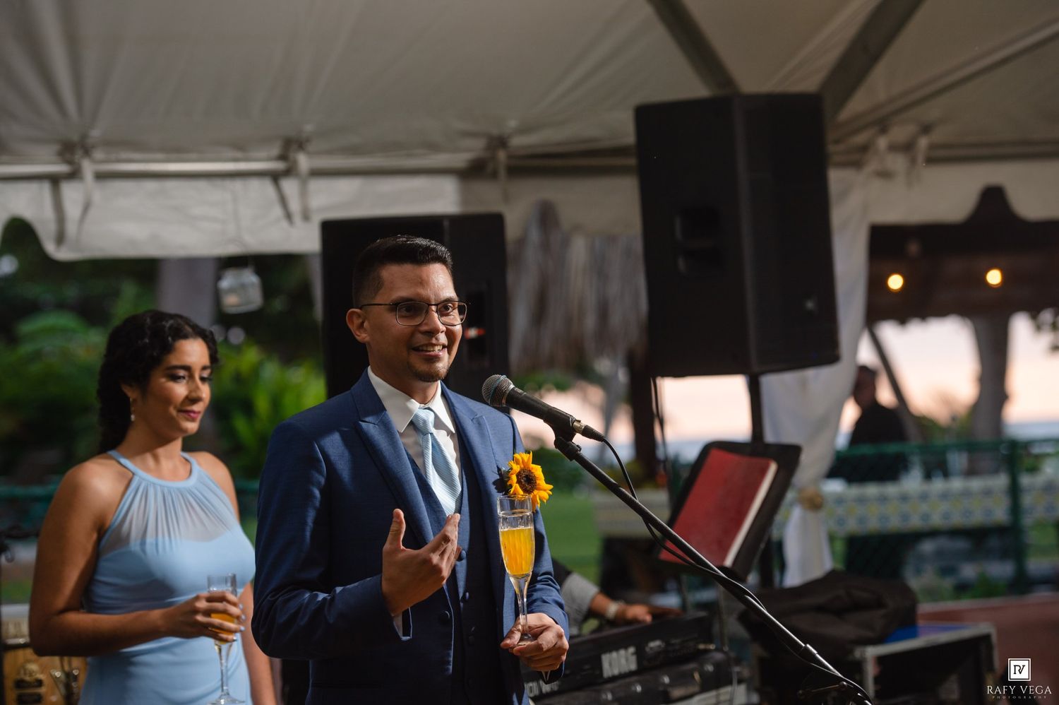 Couple in formal attire giving a speech at an outdoor wedding reception under a white tent.