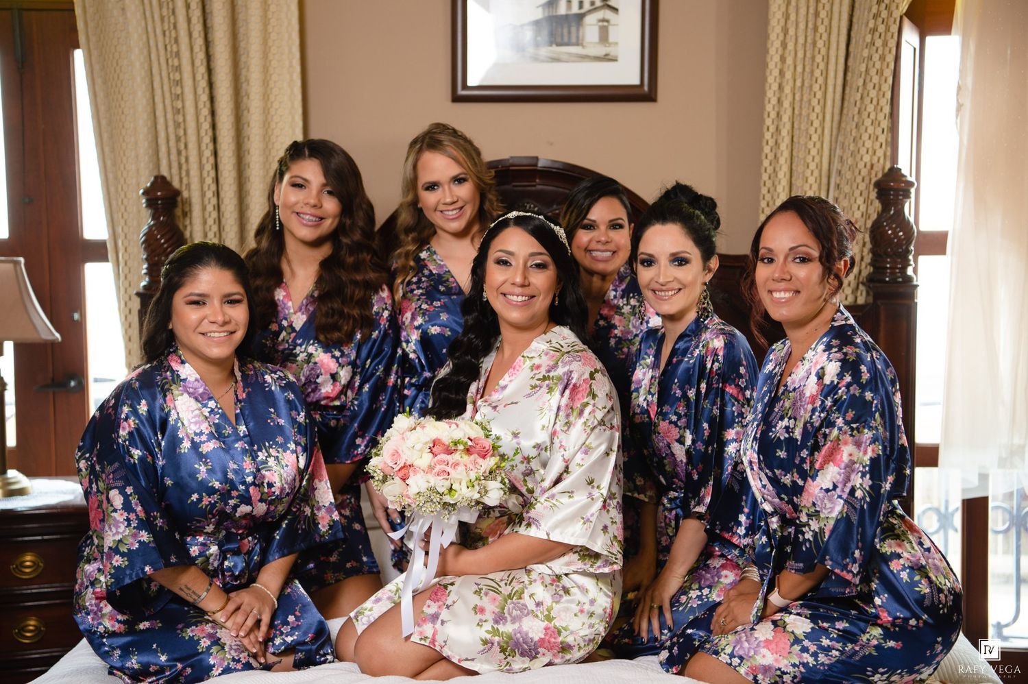 Bride and six bridesmaids wearing floral satin robes pose together smiling in a bedroom before the wedding ceremony.