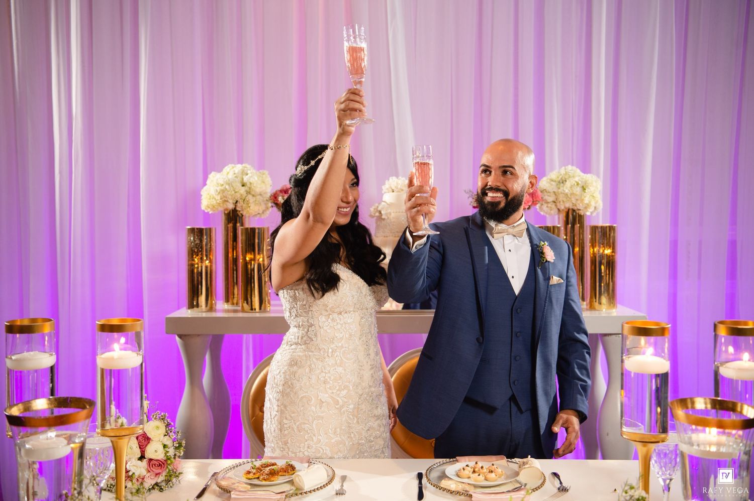 Bride and groom raising champagne glasses in a toast at their elegant wedding reception with gold decor.