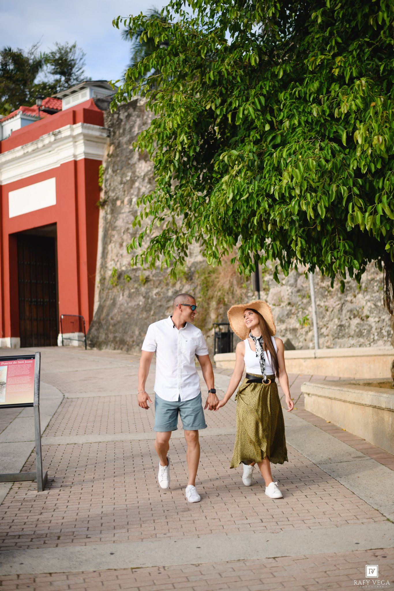 A couple walks hand-in-hand along a brick path near a red historic building, surrounded by lush green trees.