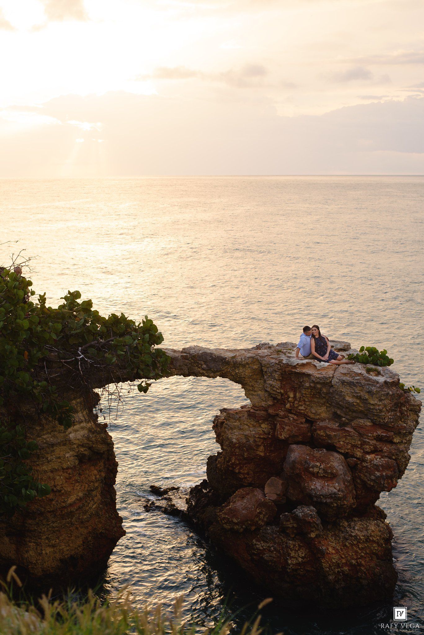 Couple sitting on rocky coastal arch at sunset overlooking calm ocean waters with golden sky.