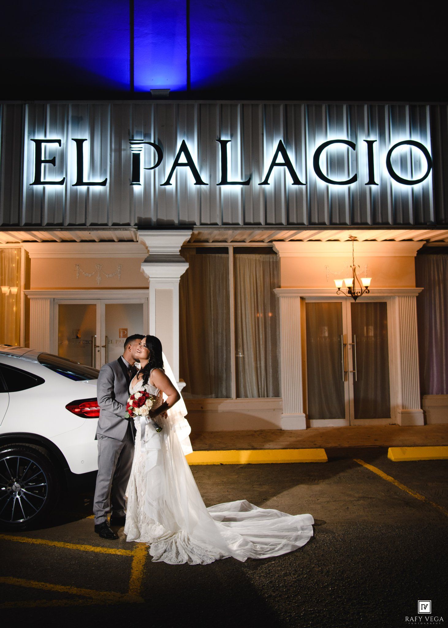 Bride and groom share a romantic kiss outside El Palacio venue at night, with blue uplighting and elegant white decor.