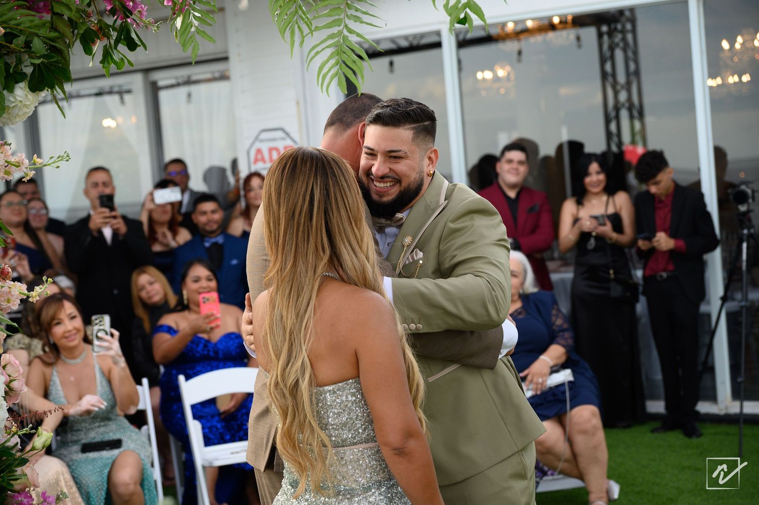 Bride in a sparkling gown and groom in an olive suit share a first dance as smiling wedding guests watch from the sides.