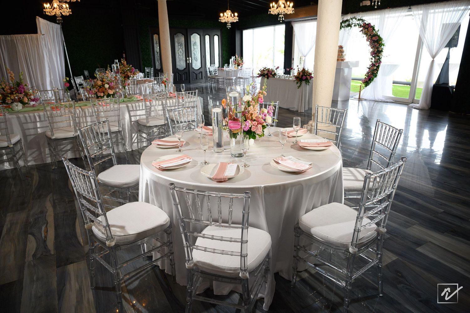 Elegant wedding reception table with a pink floral centerpiece, white linens and clear acrylic chairs in a bright, airy banquet hall.
