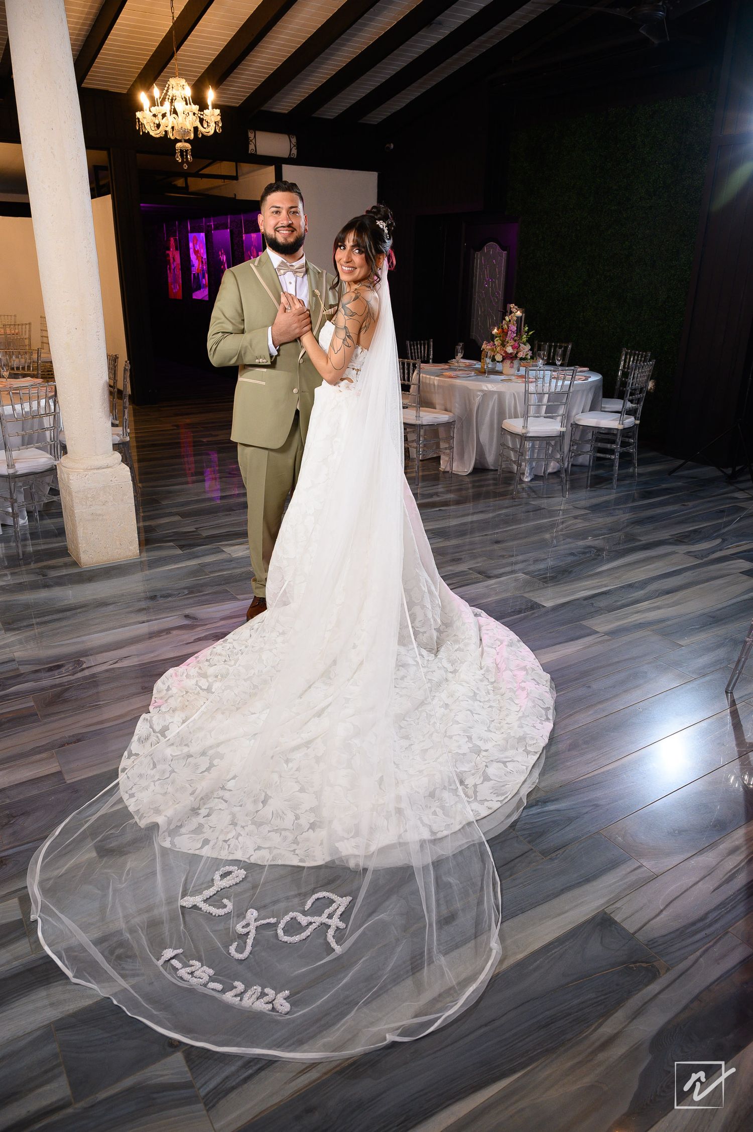 Bride in a lace wedding gown with a long embroidered veil and a groom in a green suit pose in an elegant reception hall.