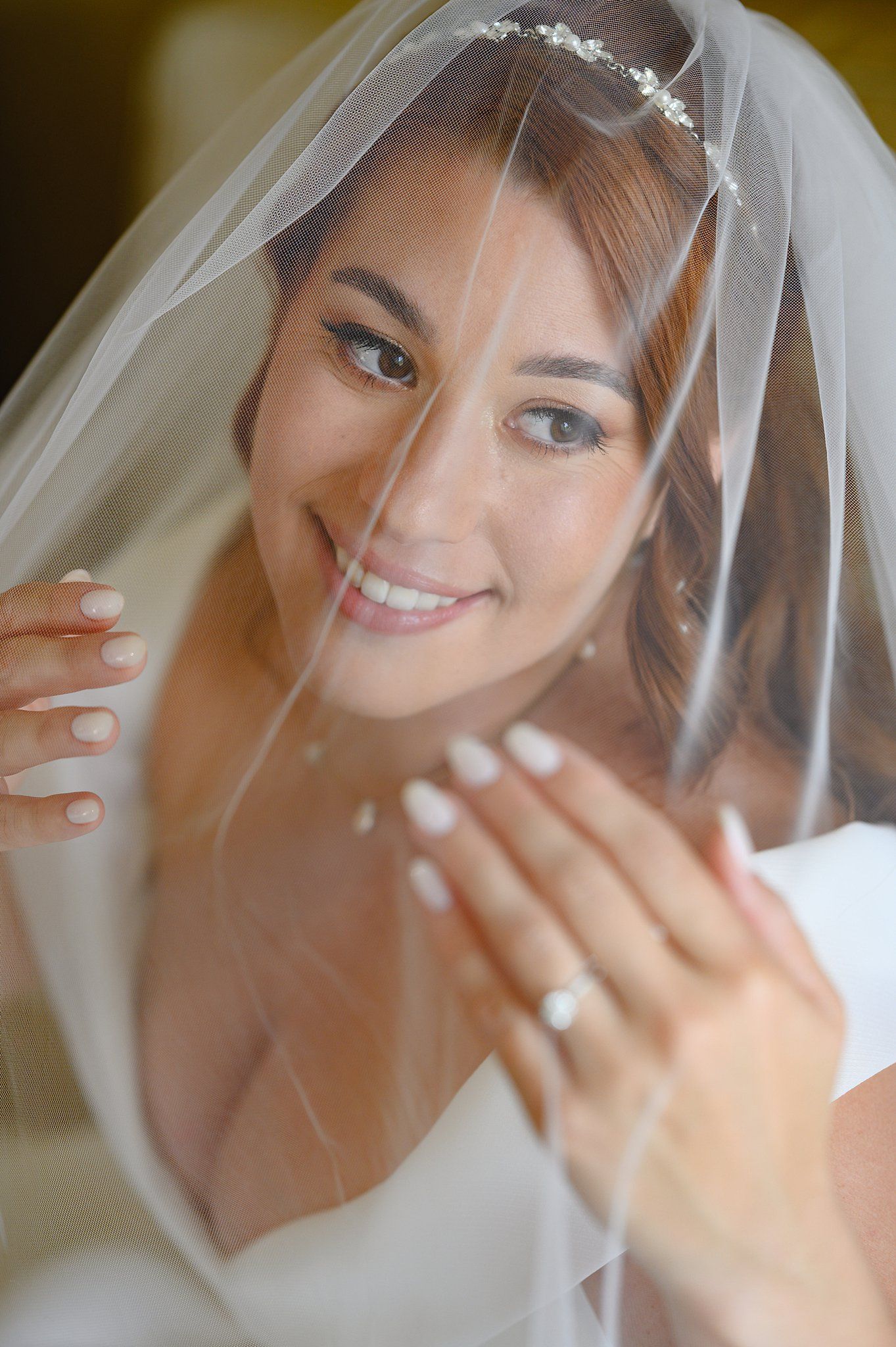 Smiling bride wearing a white veil and tiara, showing off her engagement ring on her wedding day.