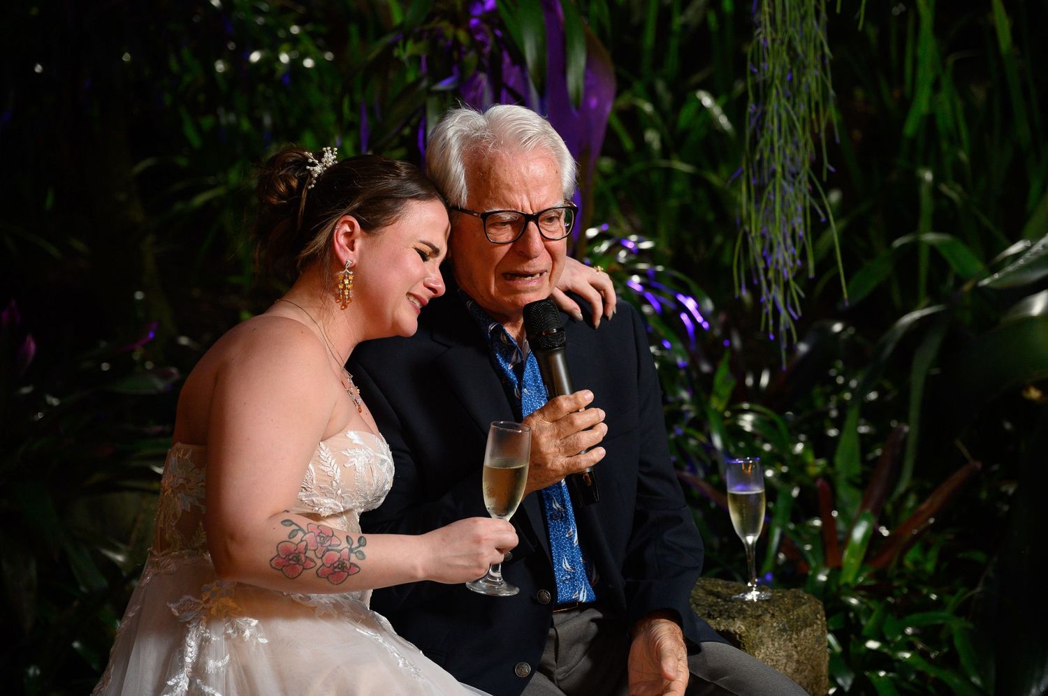 Bride in white gown shares champagne toast with elderly man in blue shirt at lush green wedding reception.