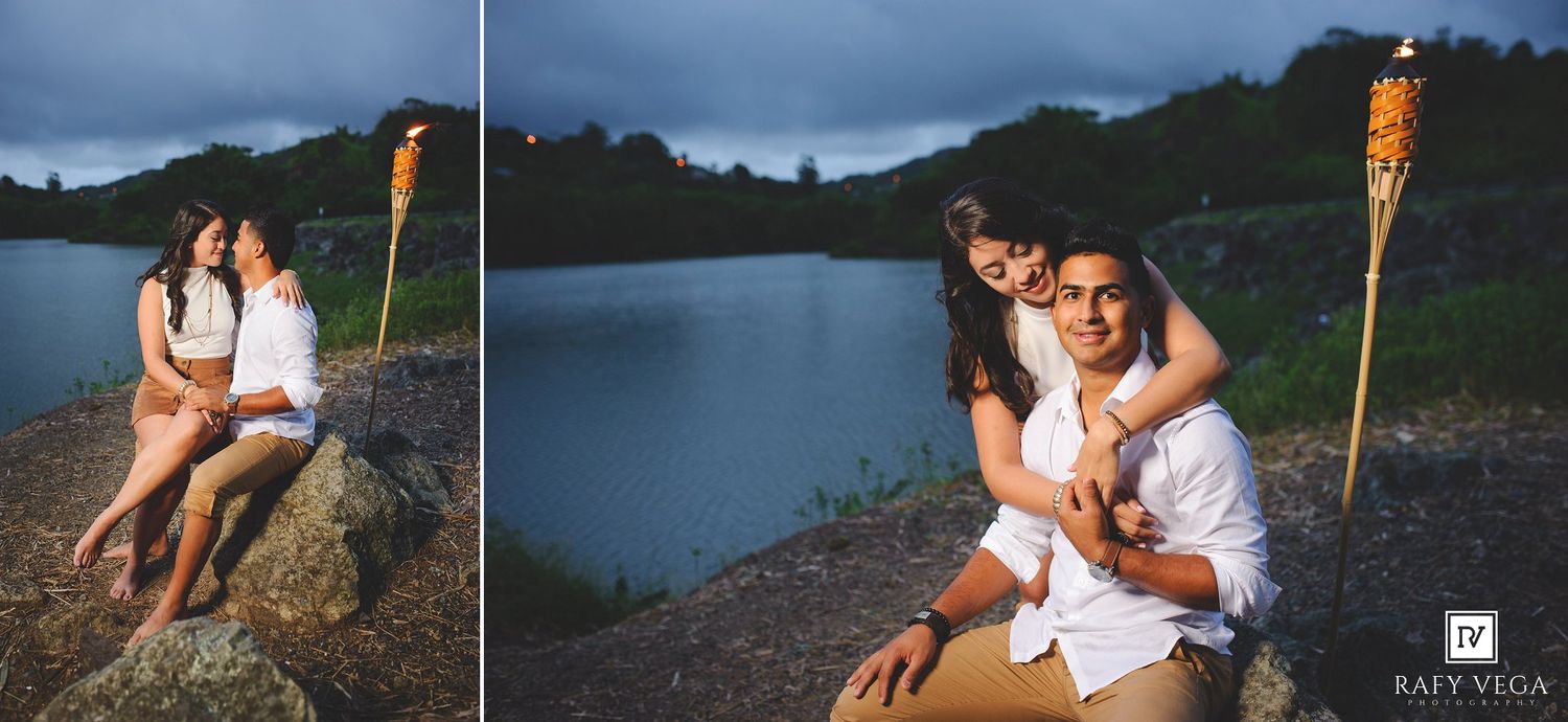 Romantic couple sitting on rocks by a lake at dusk with tiki torches, sharing an intimate embrace outdoors.