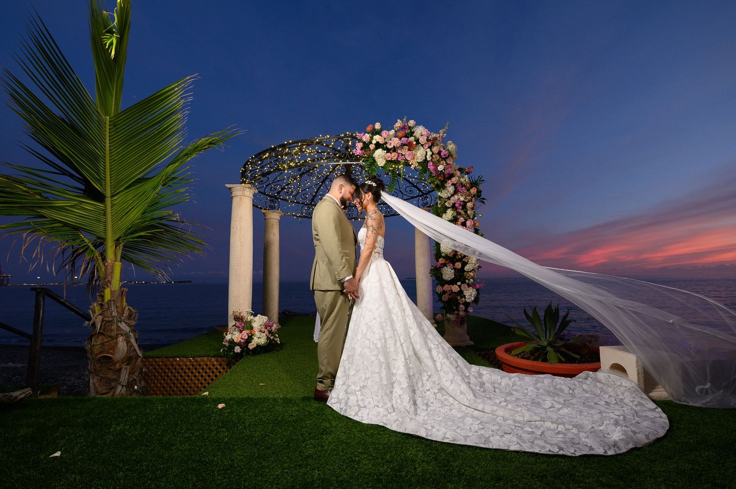 Bride and groom kissing under a floral arch at sunset with a flowing veil, oceanview wedding ceremony.