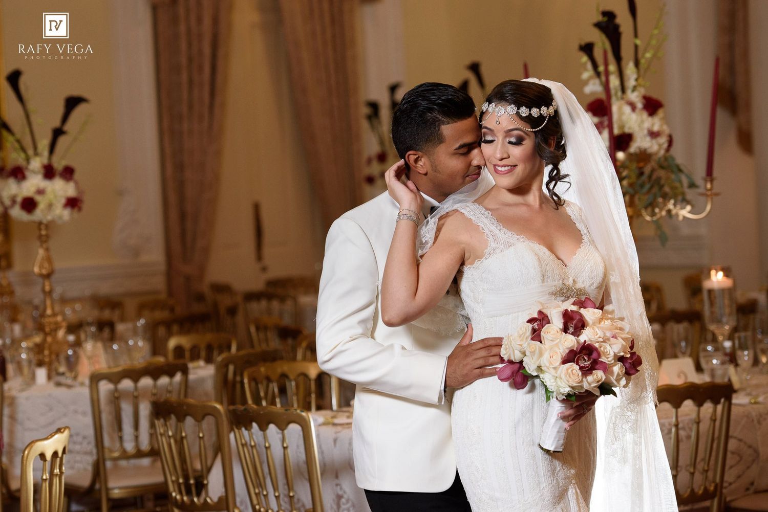 Elegant wedding couple embracing in a grand ballroom, bride in white gown holding bouquet, groom in white suit.