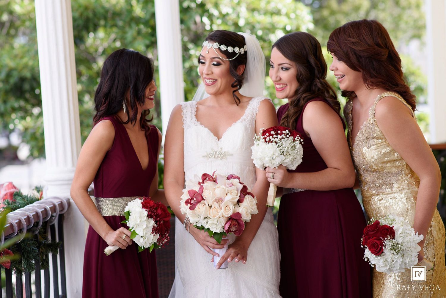 Bride in white gown with bridesmaids in burgundy and gold dresses holding bouquets on a white porch.