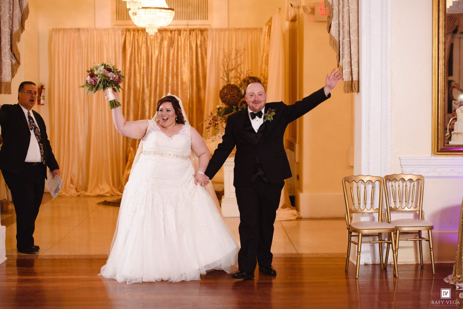 Bride and groom make grand entrance at elegant wedding reception, holding hands and waving joyfully in a gold-accented ballroom.