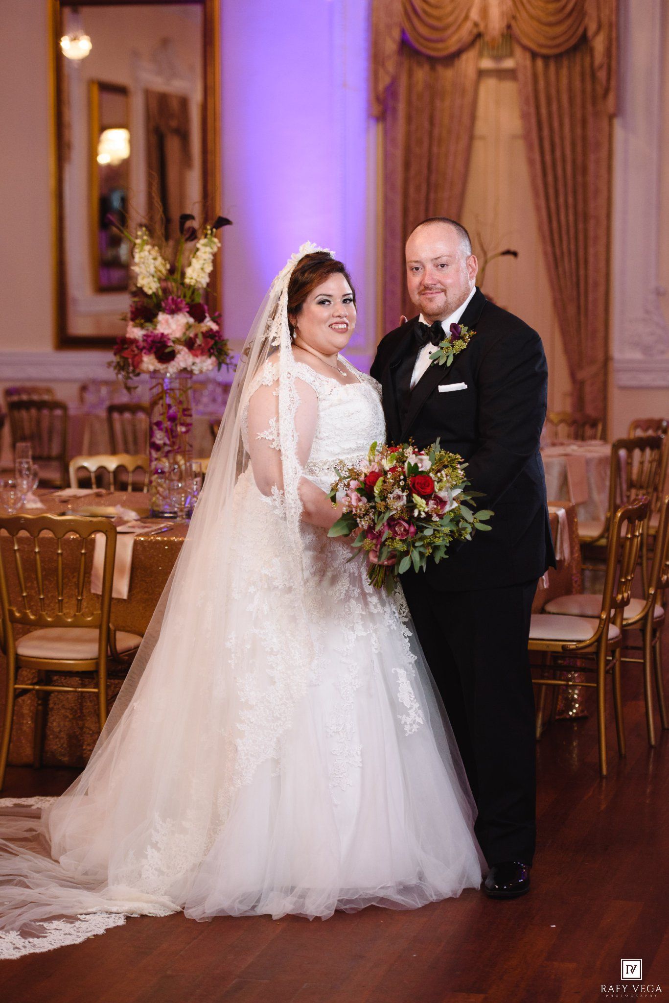 Smiling bride in lace gown with veil and groom in tuxedo pose together holding a red and green bridal bouquet.