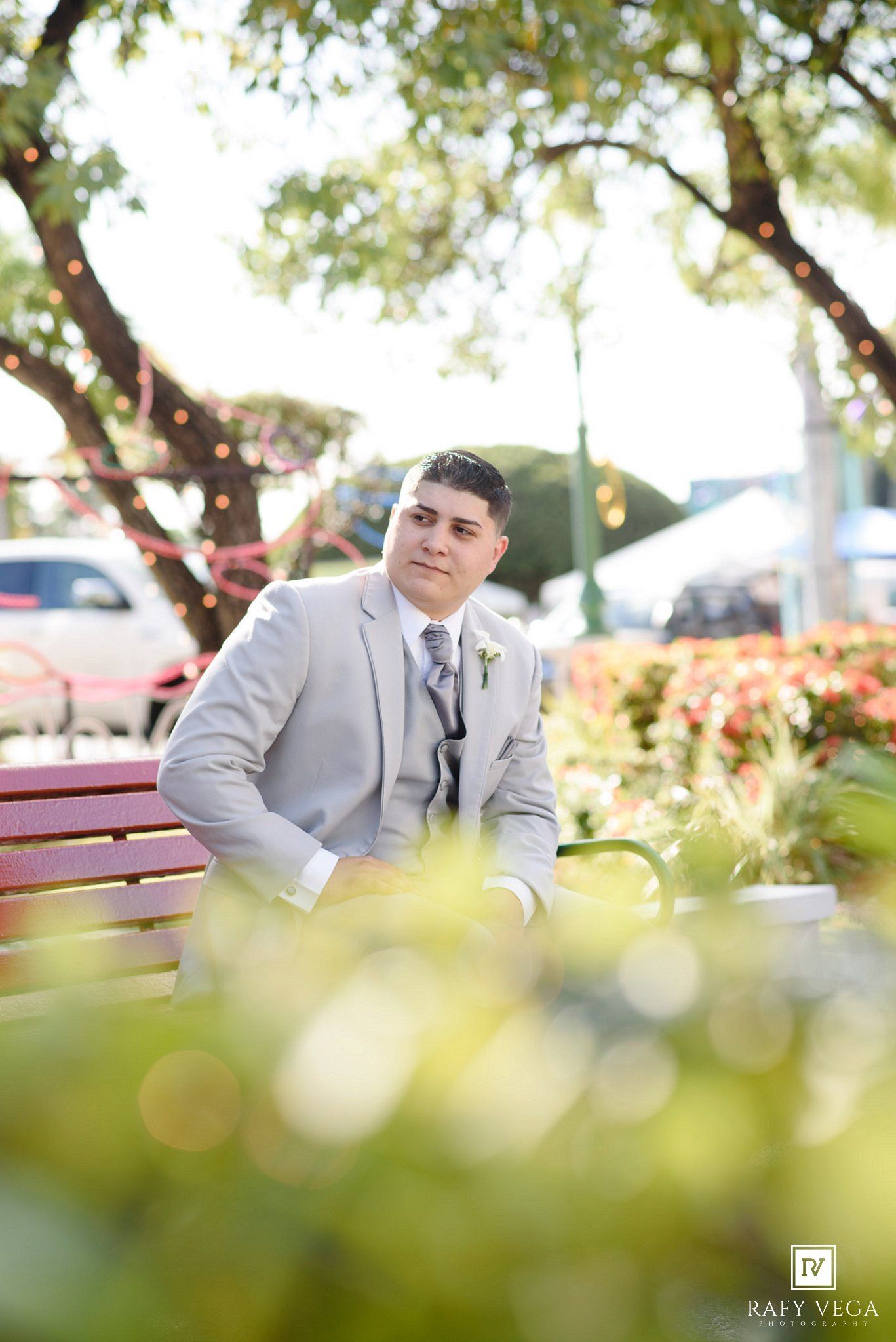 Groom in light gray suit sitting on a red bench surrounded by lush greenery at an outdoor wedding venue.