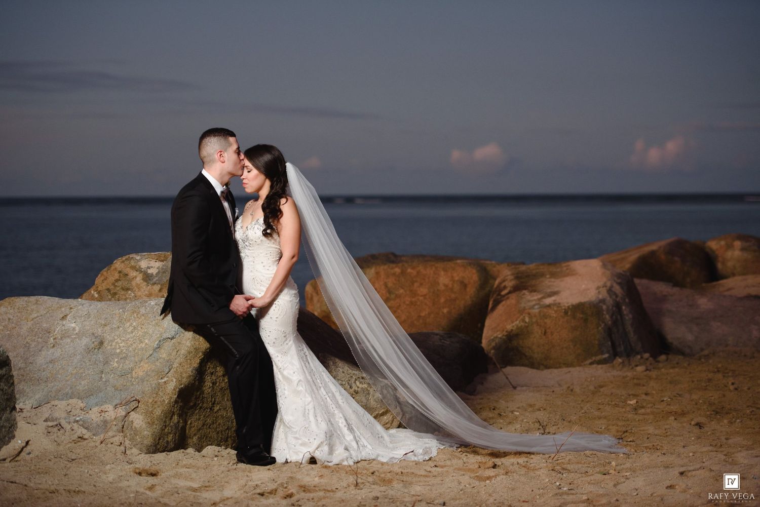 Bride and groom share a romantic kiss on a sandy beach at sunset, with rocky jetty and calm ocean in the background.