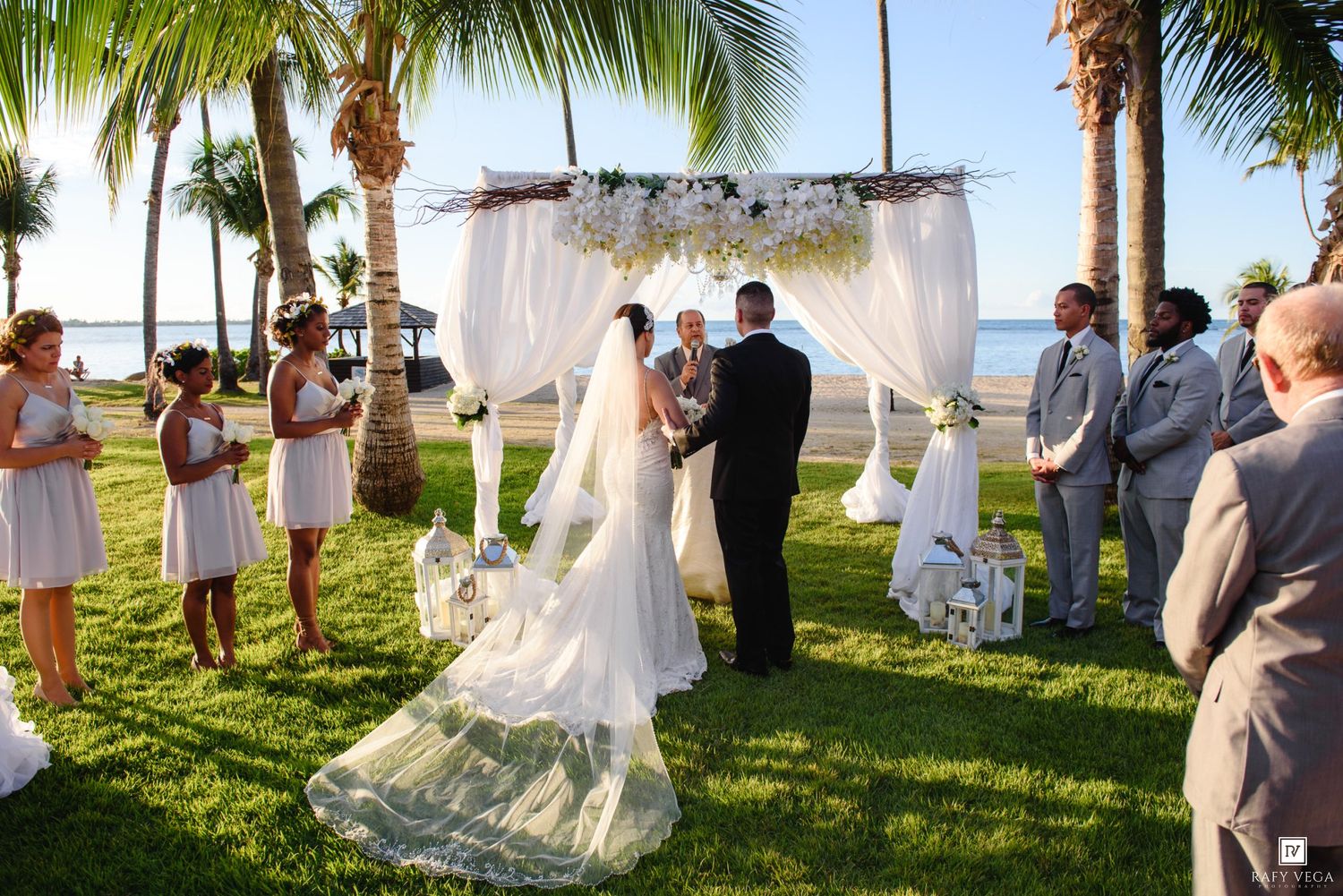 Outdoor beach wedding ceremony under white floral arch with palm trees, bride in gown, groom, and wedding party on green lawn.