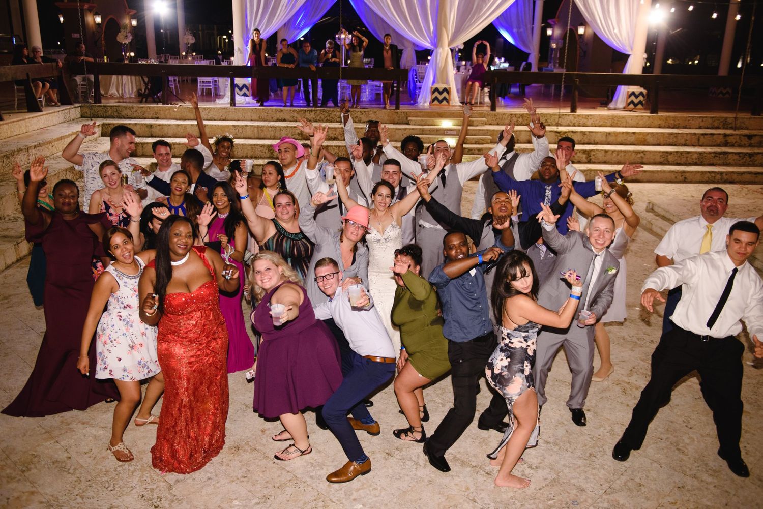 A large group of wedding guests celebrating and posing together at an outdoor evening reception venue.