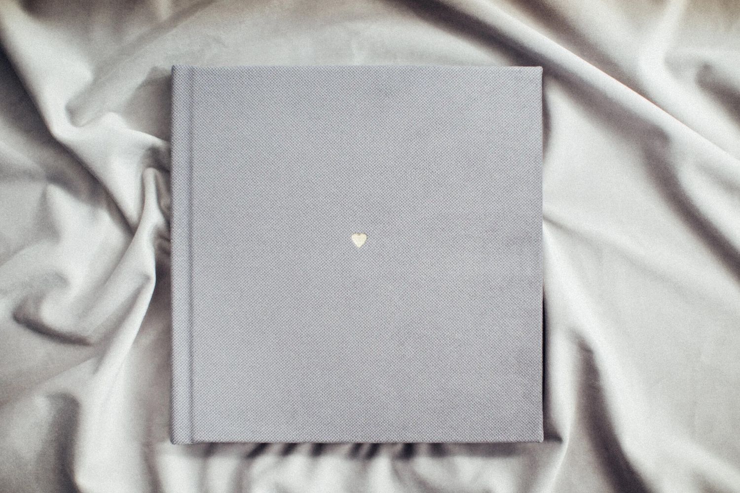 Minimalist gray hardcover wedding photo album with small gold heart emblem on silk fabric.