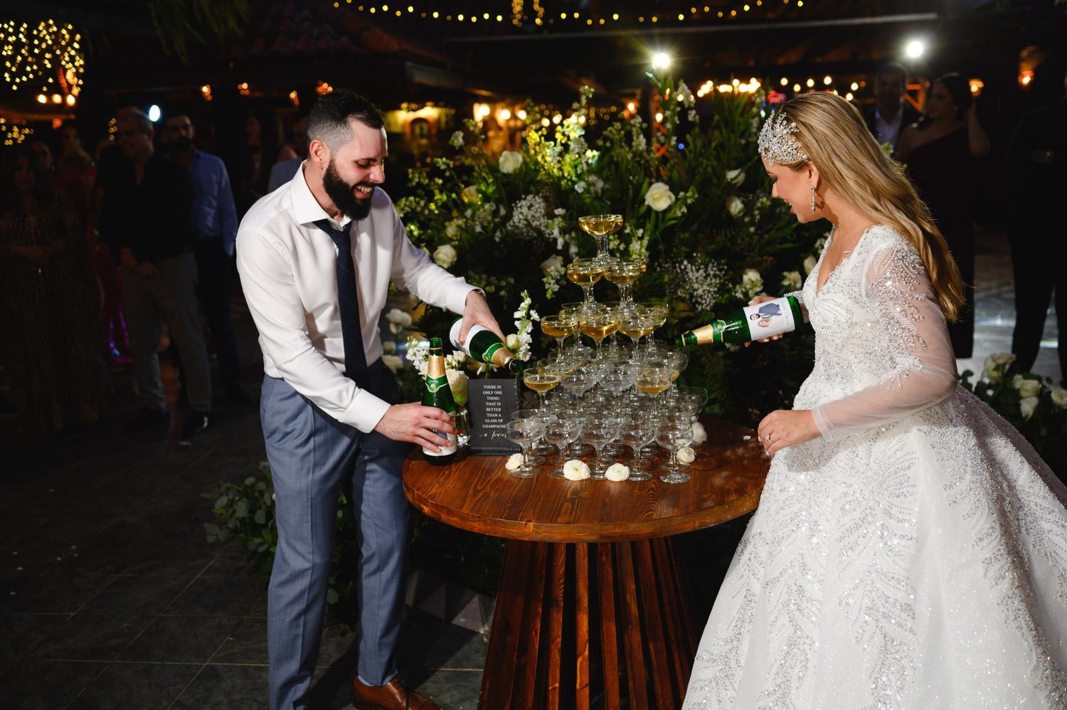 Newlyweds pour champagne tower together at elegant outdoor wedding reception at night.