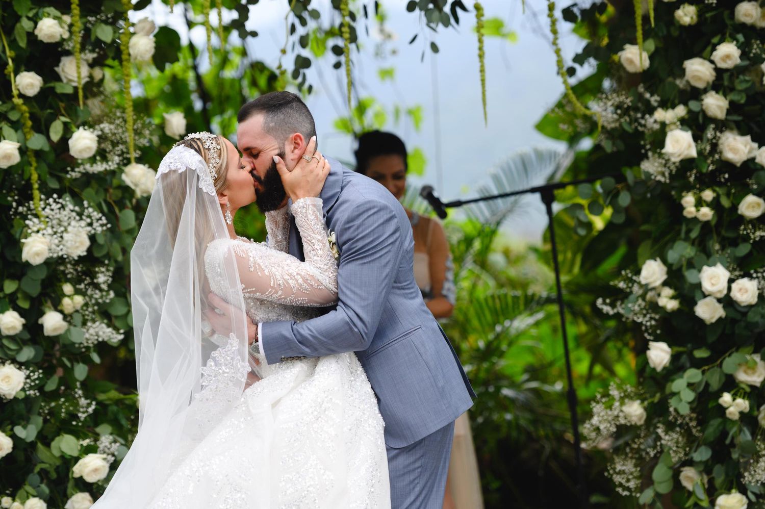 Bride and groom share first kiss at outdoor wedding ceremony beneath lush floral arch with white roses and greenery.
