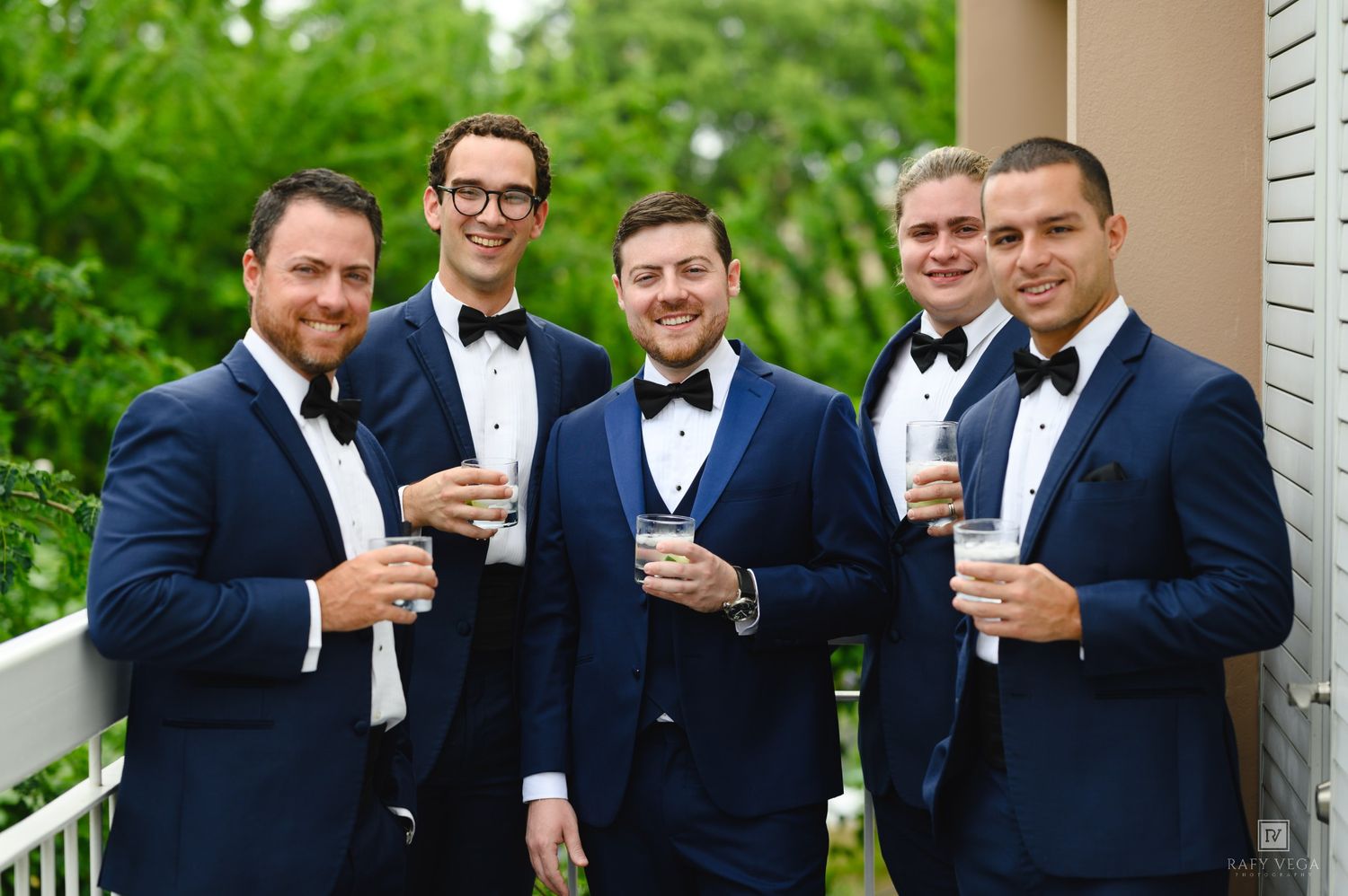 Five groomsmen in navy blue tuxedos with bow ties holding drinks and smiling outdoors.