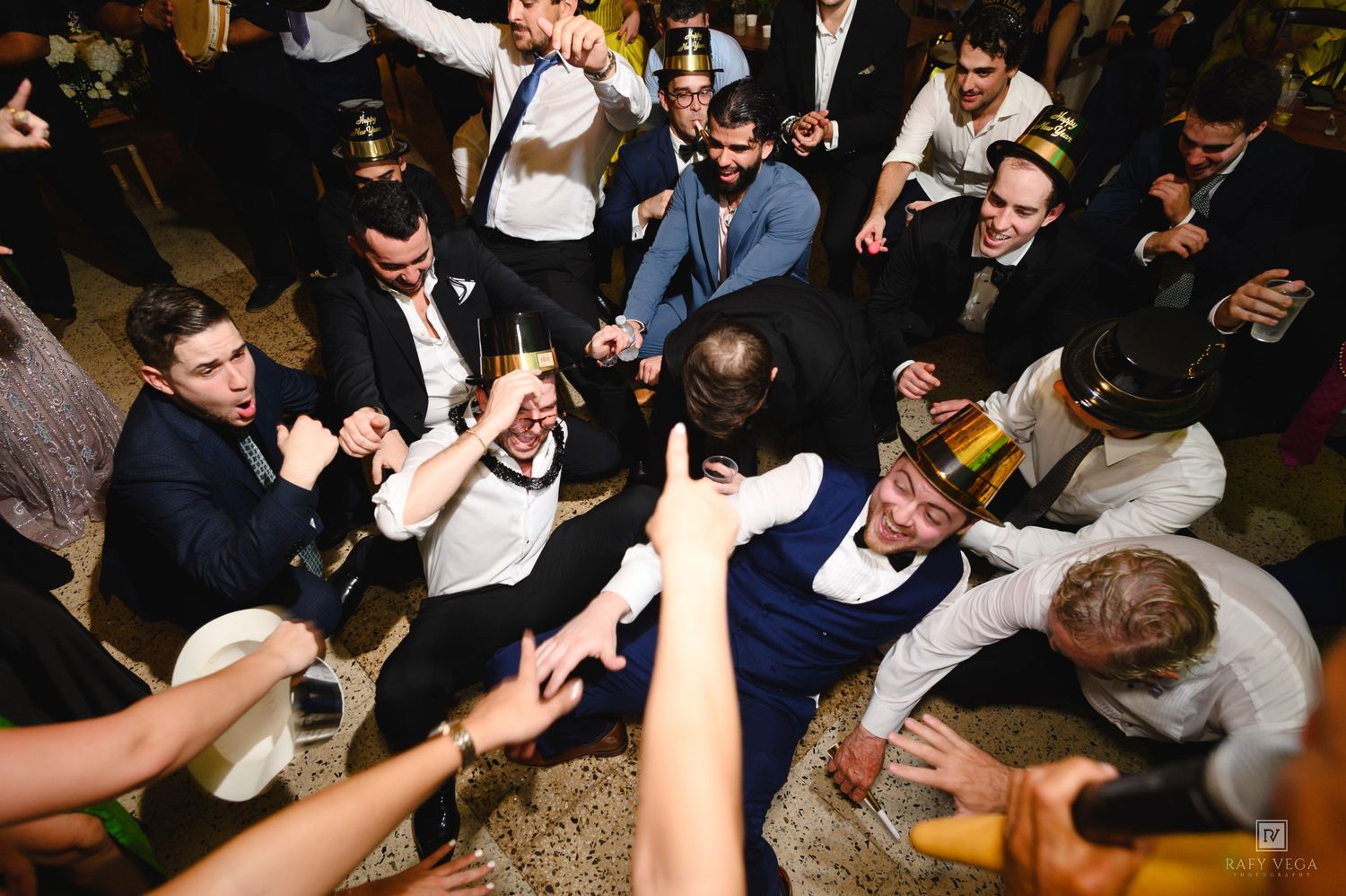 Energetic wedding reception dance floor with men in suits celebrating, raising glasses, and dancing together at a party.