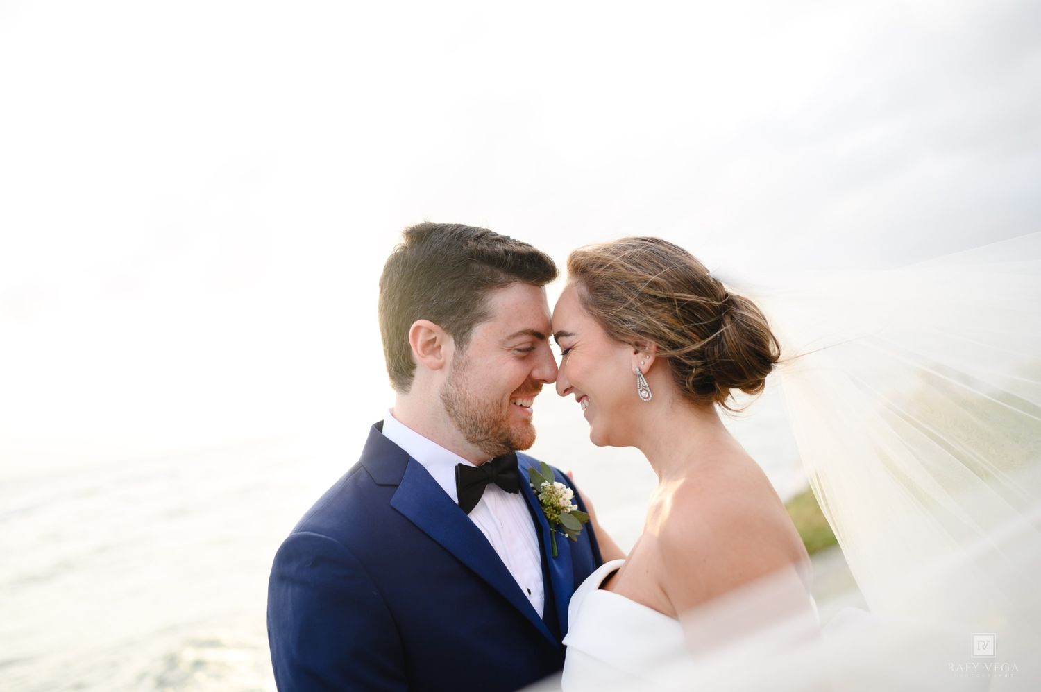 Bride and groom touching foreheads romantically outdoors, groom in navy tuxedo, bride in white gown with flowing veil.