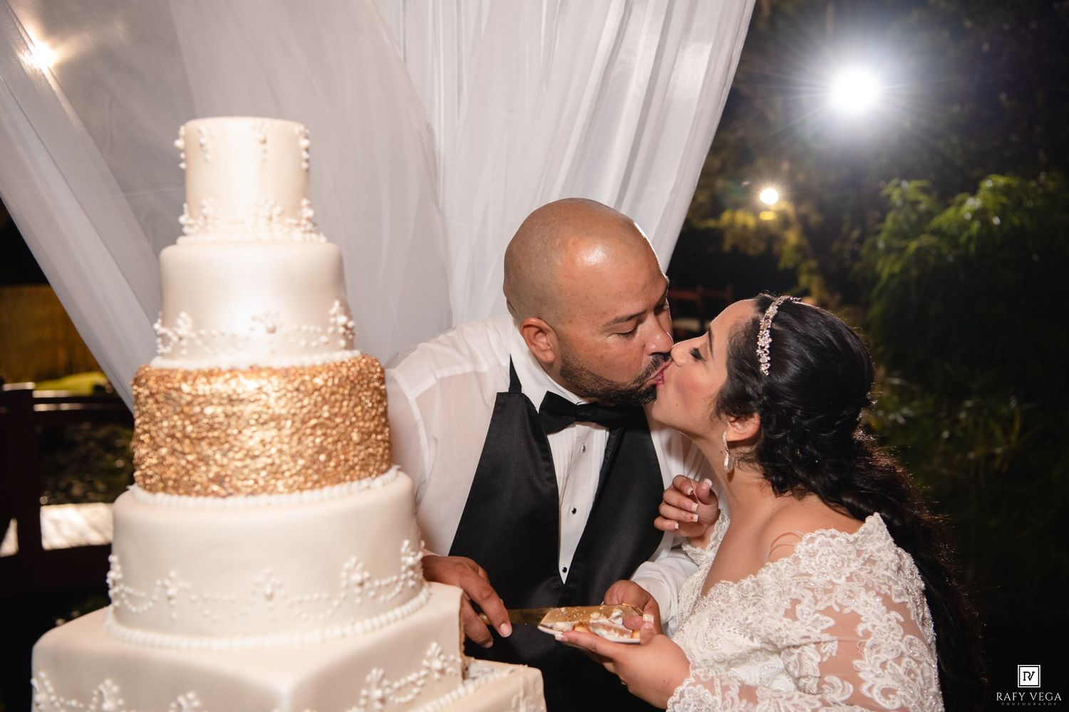 Newlyweds share a kiss while cutting their elegant four-tier white and gold wedding cake at their outdoor reception.
