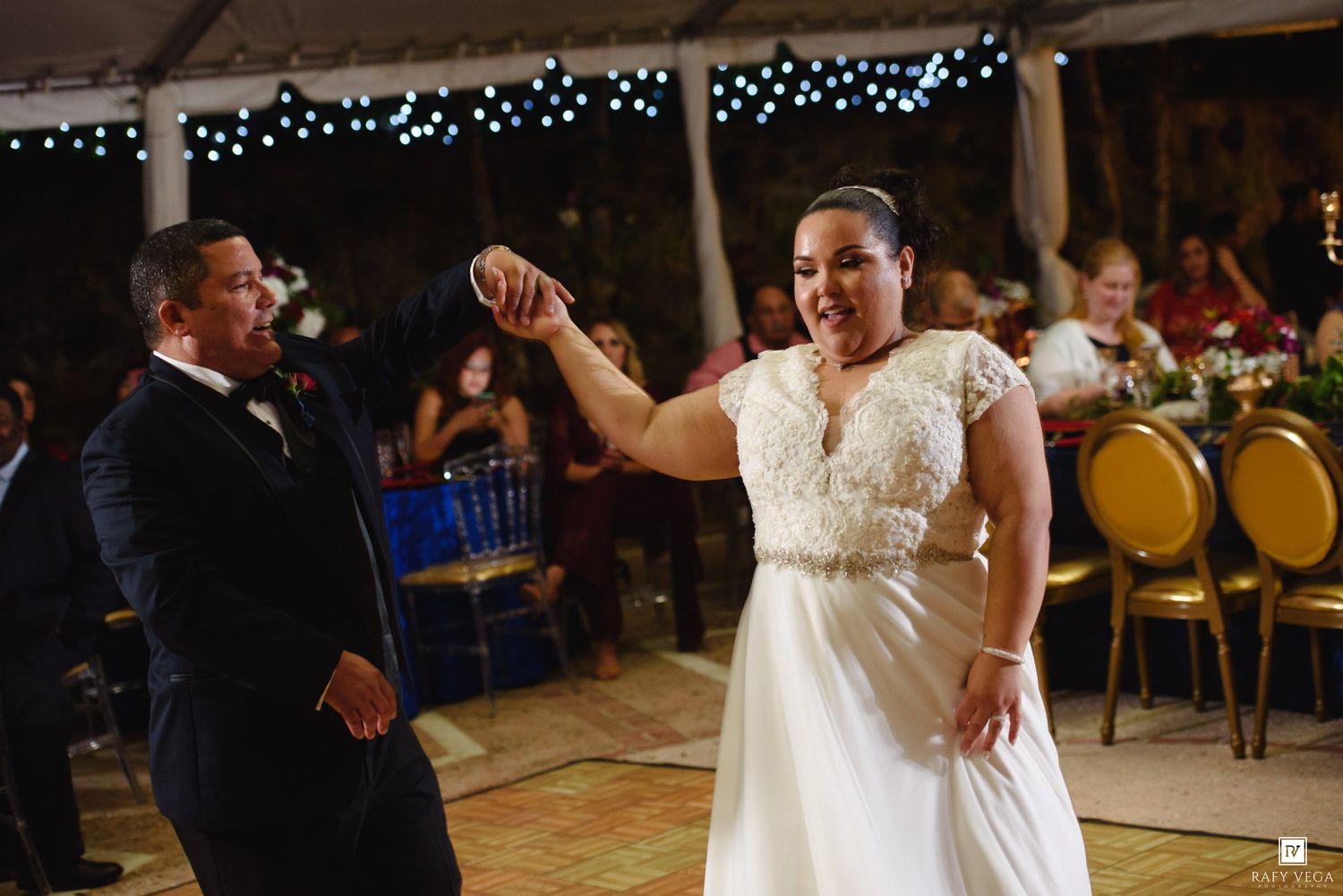 Bride in lace gown points at groom in navy suit during first dance at elegant outdoor wedding reception.