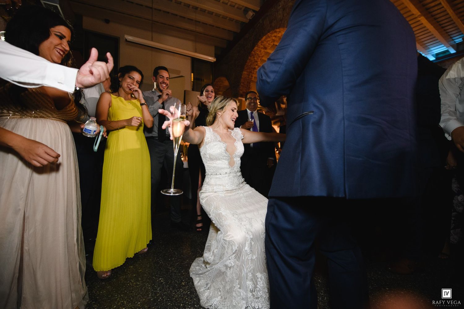 Bride in lace wedding dress dancing joyfully with guests holding sparklers at elegant indoor wedding reception.