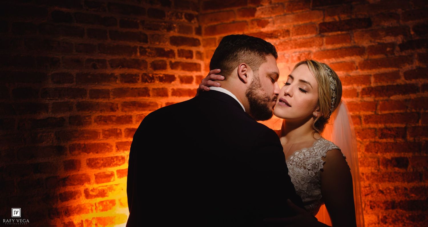 A bride and groom share a romantic moment against a warm amber-lit brick wall at their wedding reception.