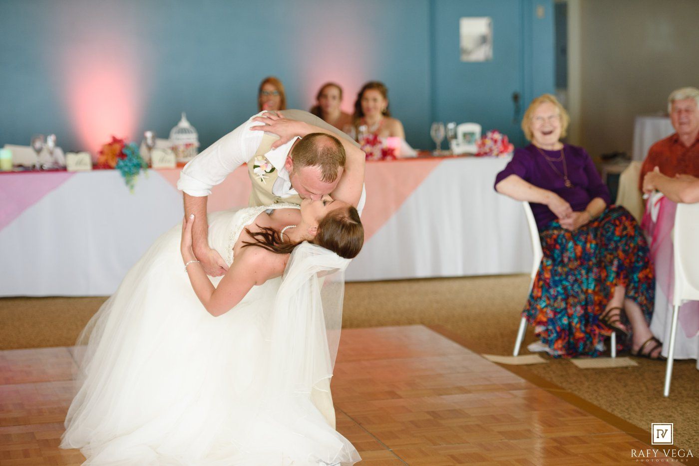 Groom dips bride for romantic kiss on dance floor at wedding reception while guests laugh and cheer.