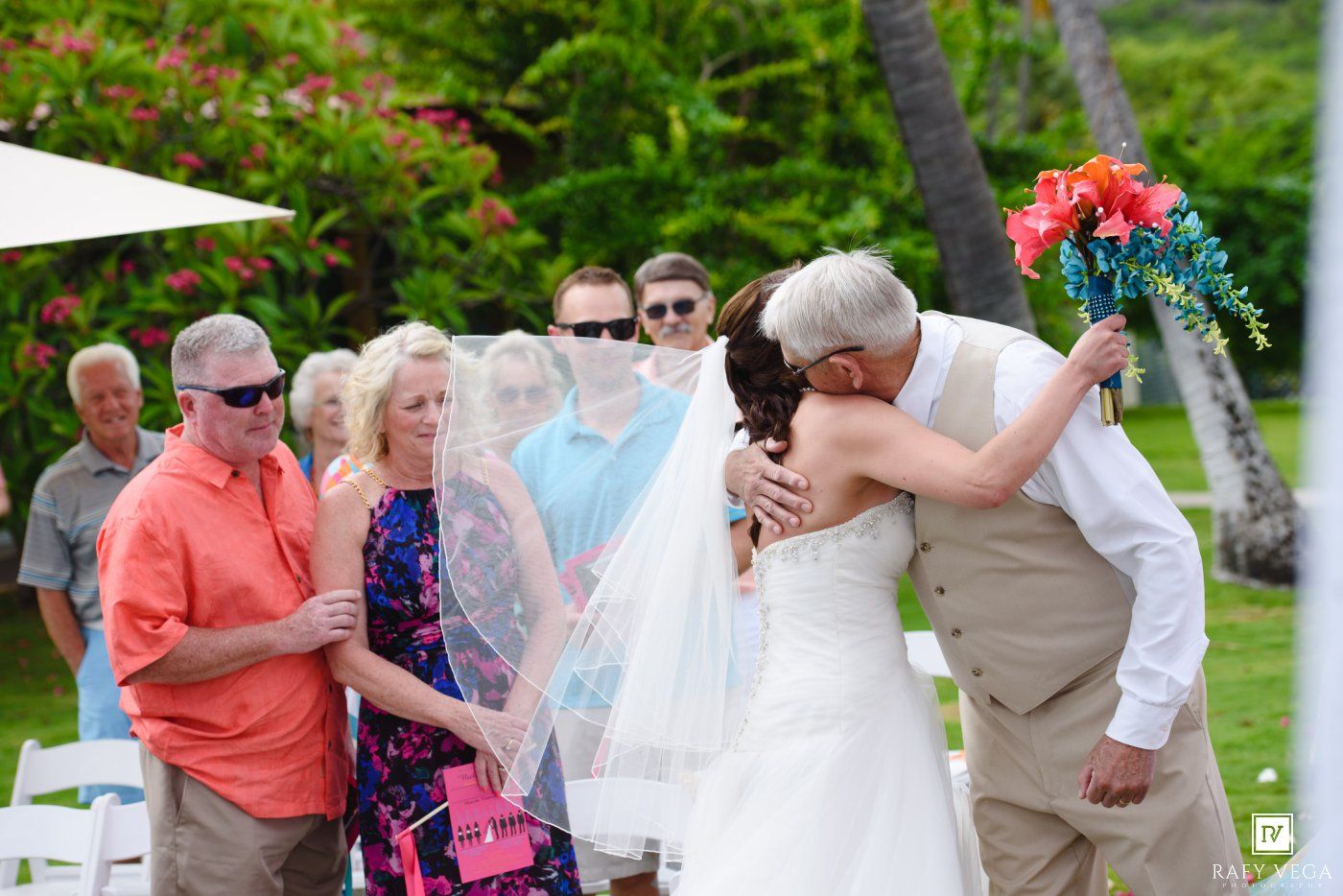 Bride embracing her father at an outdoor wedding ceremony with guests watching, holding a vibrant tropical bouquet.