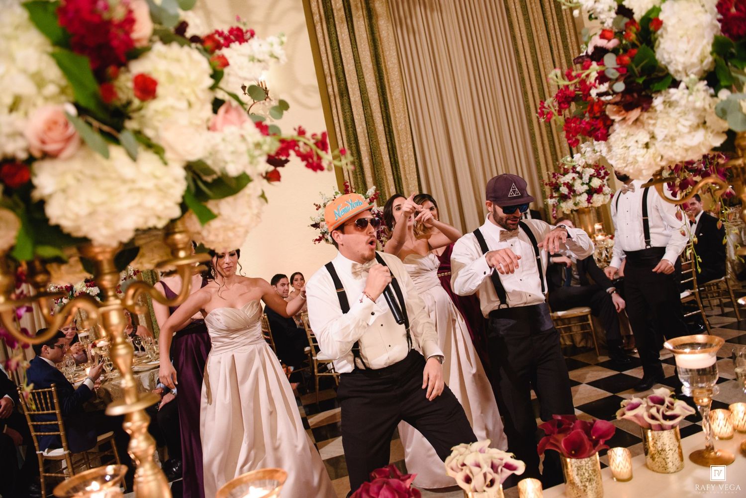 Wedding reception guests dance joyfully amid lush floral arrangements of white and red flowers on golden stands.