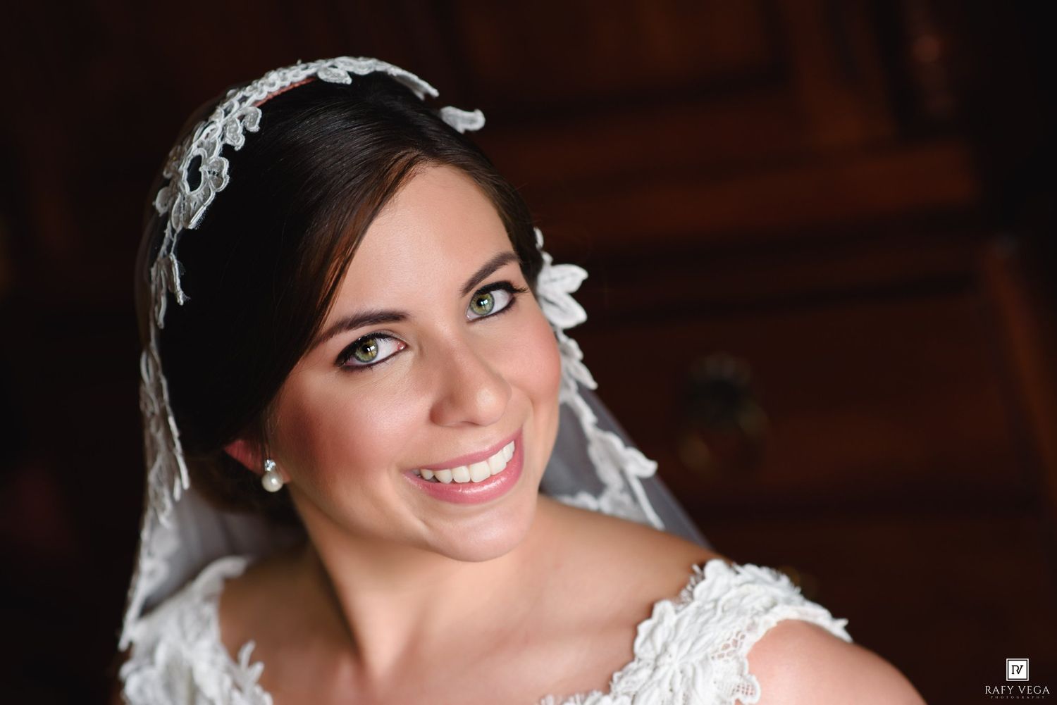 Beautiful bride with green eyes wearing lace headpiece and veil, smiling elegantly on her wedding day.