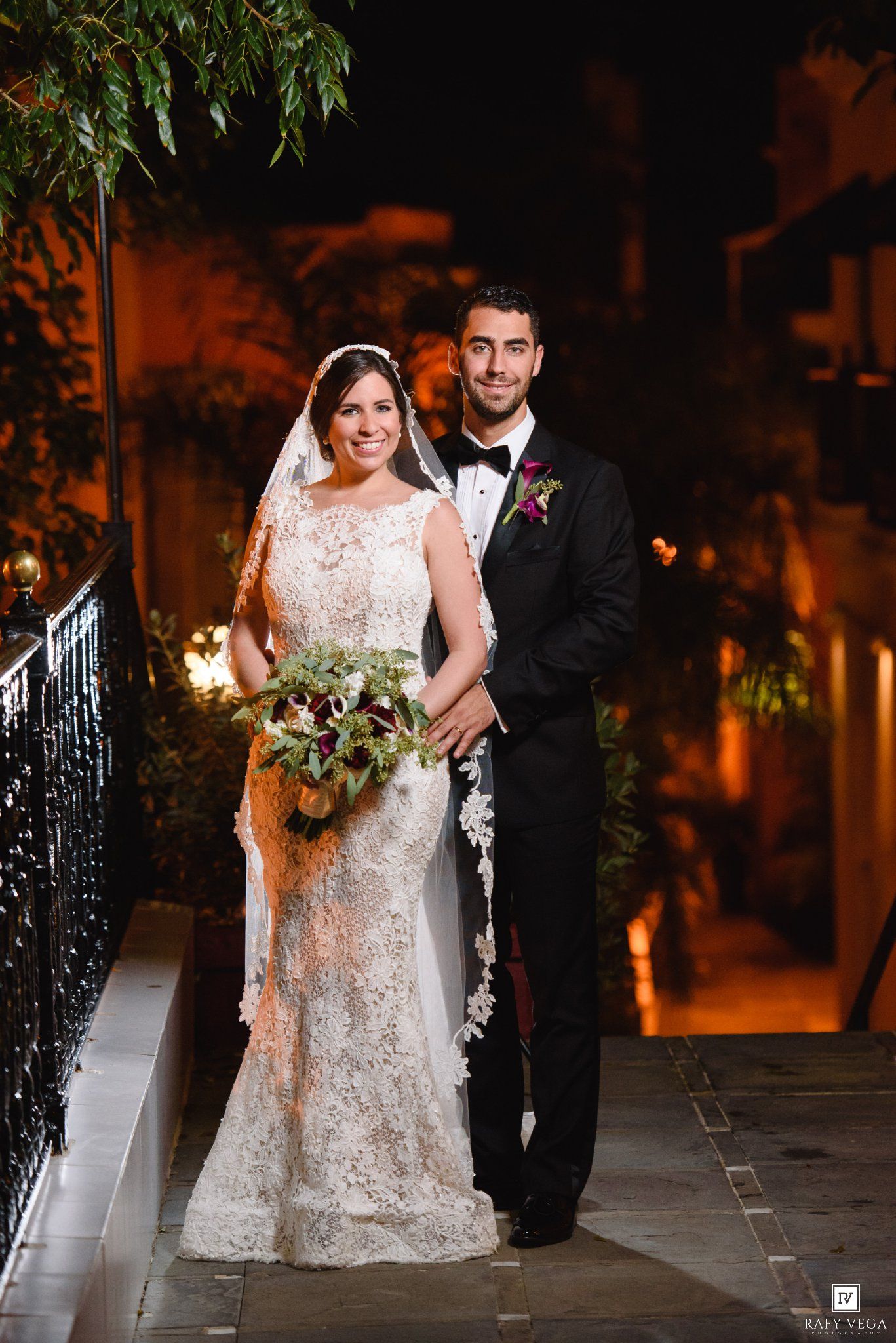 Bride in lace gown with veil and groom in black tuxedo pose together outdoors at night holding a bouquet.