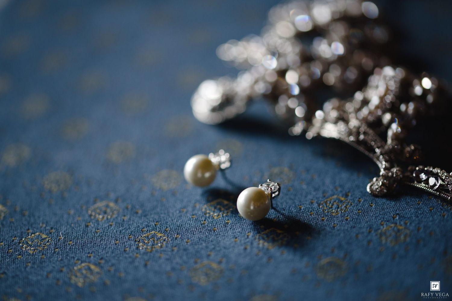 Pearl stud earrings resting on blue fabric beside silver jewelry chain in elegant close-up shot.