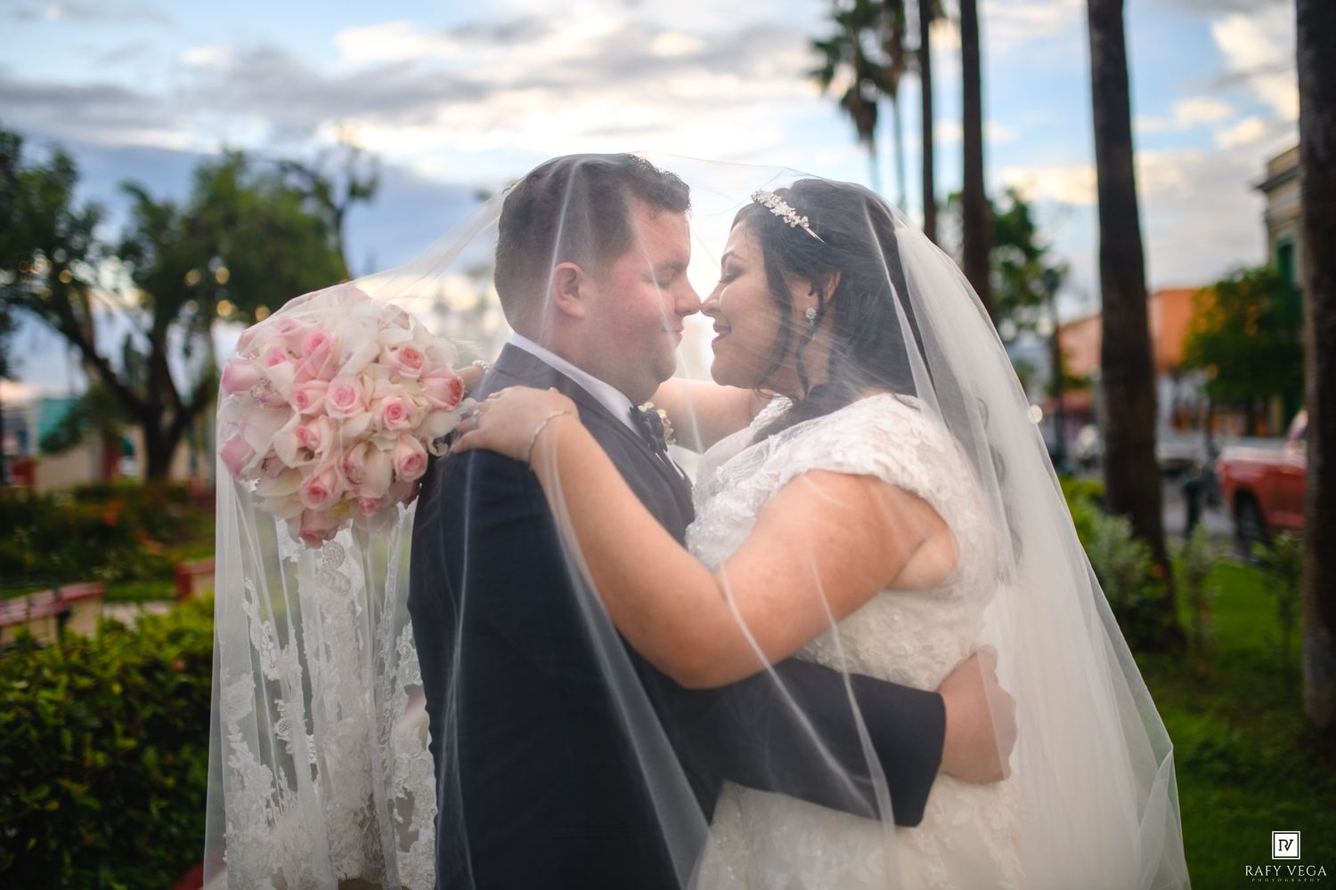 A bride and groom share a romantic moment outdoors, the bride wearing a lace gown and veil, holding a pink bouquet.