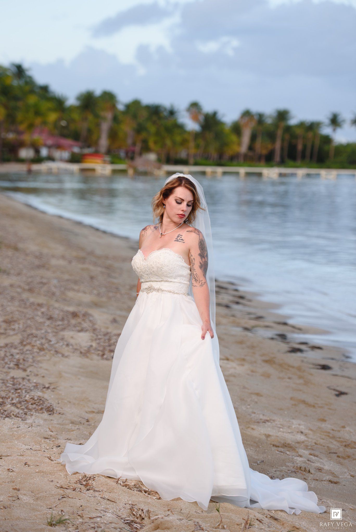 Bride in white lace wedding gown and veil poses on tropical beach shoreline with palm trees in background.