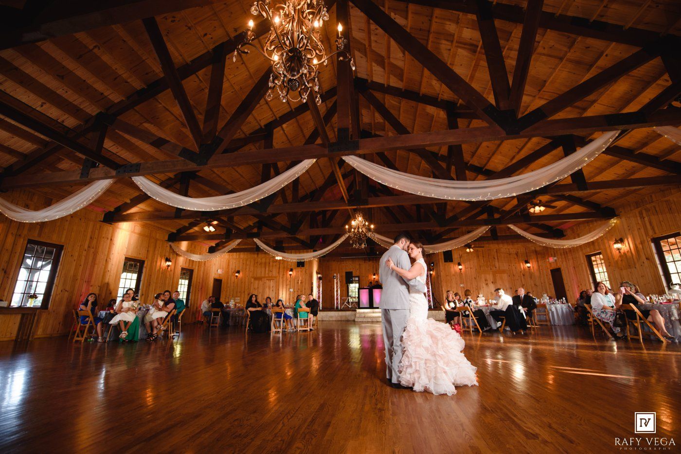 Bride and groom share first dance in elegant rustic barn wedding venue with draped white fabric and chandelier lighting.