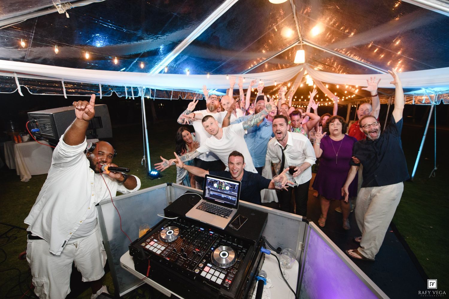DJ and excited wedding guests celebrating under a festive tent with string lights at an outdoor evening reception.