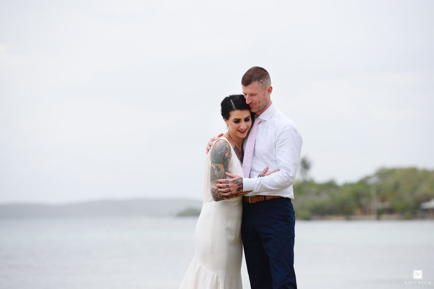 Couple embracing on a beach during their wedding, wearing white dress and blue pants, with ocean and trees in the background.