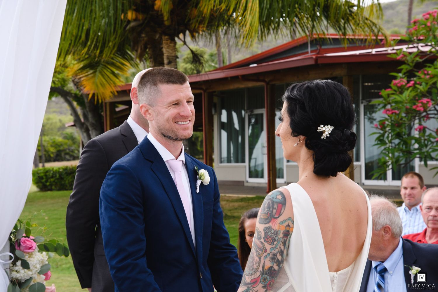 Groom in navy suit smiles at tattooed bride in white dress during outdoor wedding ceremony with tropical greenery.