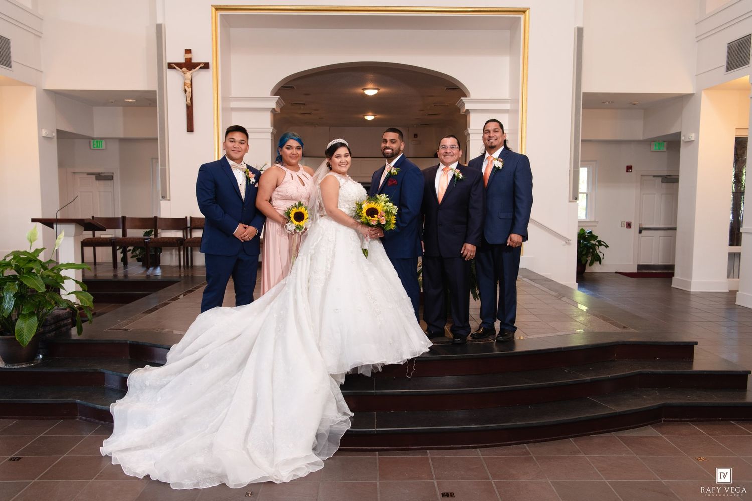 Wedding party poses in church with bride in white gown, groom, and guests holding sunflowers near altar.