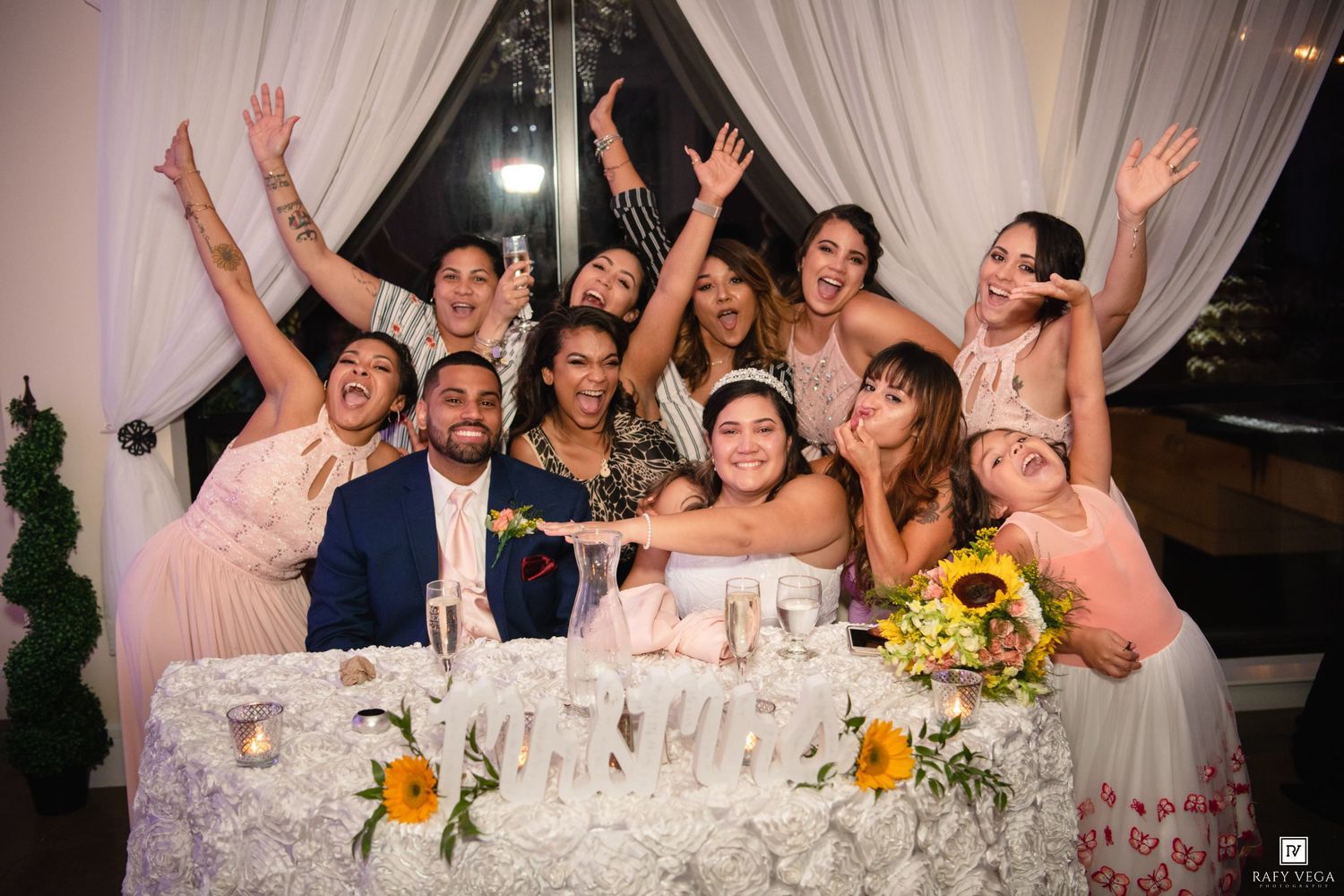 Wedding party celebrating joyfully at reception table with sunflowers, bridesmaids raising hands in excitement around bride and groom.